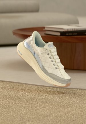 Baskets blanches matelassées avec des accents argentés et une semelle épaisse flottant au-dessus d'un tapis beige dans un salon moderne.