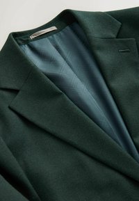 Donkergroene blazer gemaakt van textiel met textuur, met een geknikte kraag en een gladde teal voering met een geometrisch patroon.