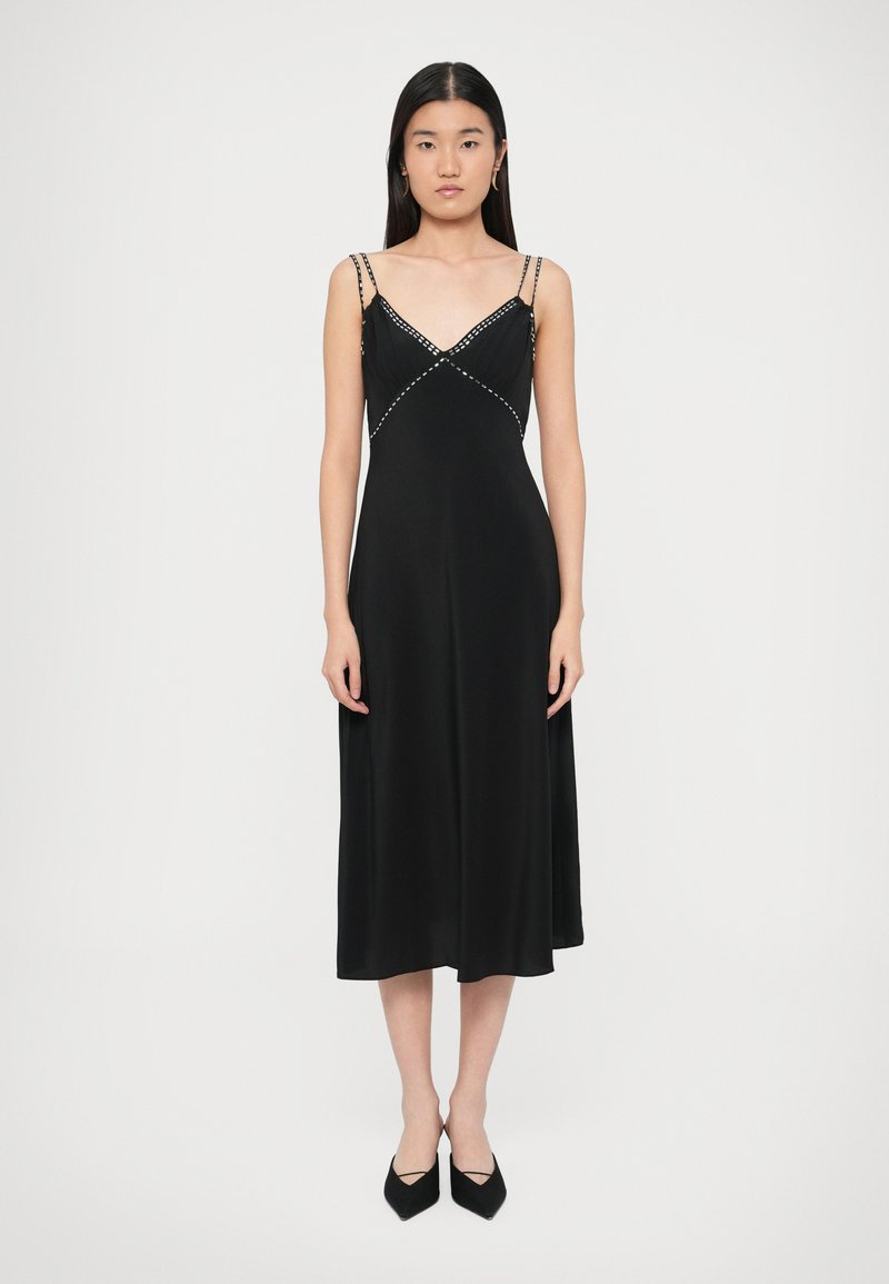 Czarna sukienka midi z dekoltem w serek, cienkimi ramiączkami i ukośnymi zdobieniami; gładka faktura; zestawiona z czarnymi butami na szpic.