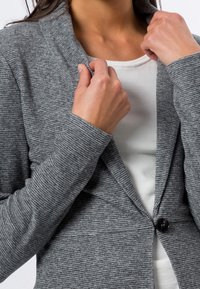 Grijze cardigan met een gestructureerde brei, sluiting met één knoop en een open kraagontwerp, gedragen over een wit shirt. Gedetailleerde mouwen zichtbaar.