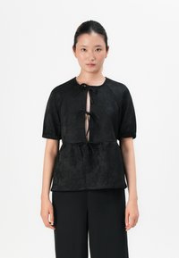 Donna con i capelli scuri raccolti indossa un blusa nera a maniche corte con tre lacci sul davanti e pantaloni neri larghi, in piedi contro uno sfondo bianco.