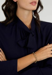 Bracciale in oro con maglie circolari e dettagli testurizzati, indossato su un polso abbronzato, abbinato a una blusa blu scuro con colletto a fiocco.