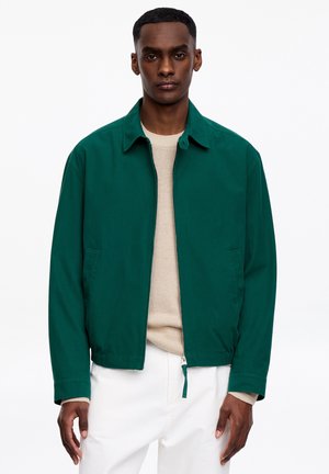 Homme portant une veste zippée vert foncé par-dessus un pull beige et un pantalon blanc, se tenant devant un fond blanc uni.