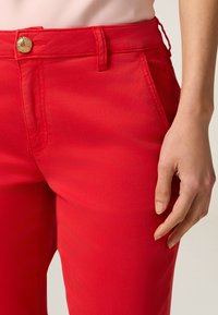 Pantaloni in cotone rosso con una texture liscia, caratterizzati da una chiusura con bottone dorato e tasche laterali. Il design include passanti per la cintura.