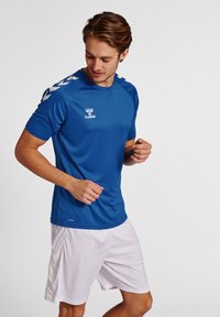 Fiatal férfi kék hummel sportpólóban és fehér rövidnadrágban, egyszínű fehér háttér előtt pózol, lefelé nézve.