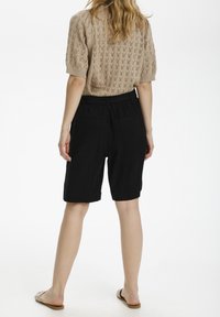 Femme de dos portant un haut en tricot beige à manches courtes, un short noir jusqu'aux genoux et des sandales slides beiges.