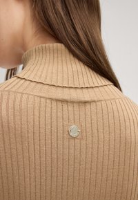 Beige gerippter Pullover mit einem umgeschlagenen Kragen und einem dekorativen Metalllogo auf der Rückseite. Die Textur wirkt weich und körpernah.