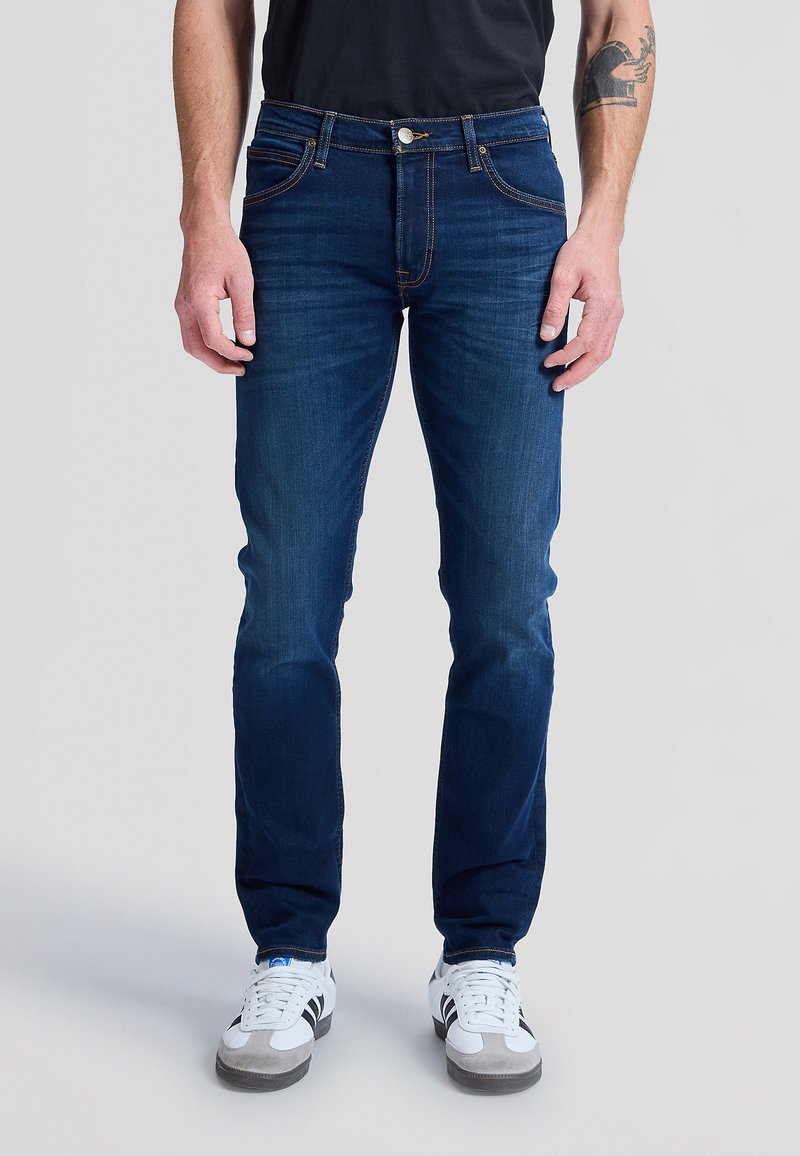 Man draagt slim-fit donkerblauwe spijkerbroek met oranje stiksels en witte sneakers, staand tegen een effen achtergrond.