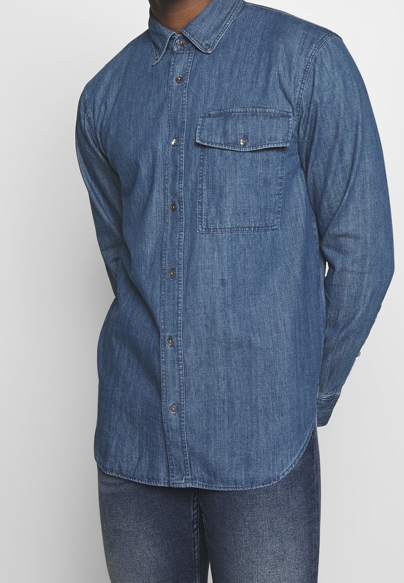 Camicia in denim con colletto a punta, maniche lunghe e una sola tasca sul petto; colore blu scuro con una finitura strutturata e un sottile motivo verticale.