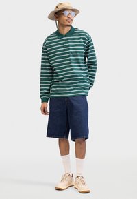 Green and white striped long-sleeve polo shirt, denim shorts, beige sneakers, white socks, and a beige bucket hat with sunglasses.