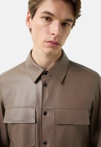 Brun læderskjorte med knaplukning, spids krave og to klaplommer. Glat tekstur med diskrete syningsdetaljer.