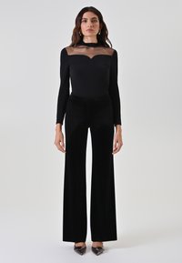 Body a manica lunga nero con scollo trasparente, abbinato a pantaloni neri a gamba larga in velluto e tacchi a punta.