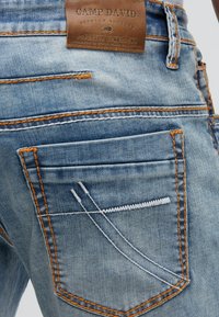 Jeans en denim bleu clair avec coutures orange, motif brodé blanc sur la poche arrière, et une étiquette en cuir marron de la marque Camp David au-dessus de la poche.