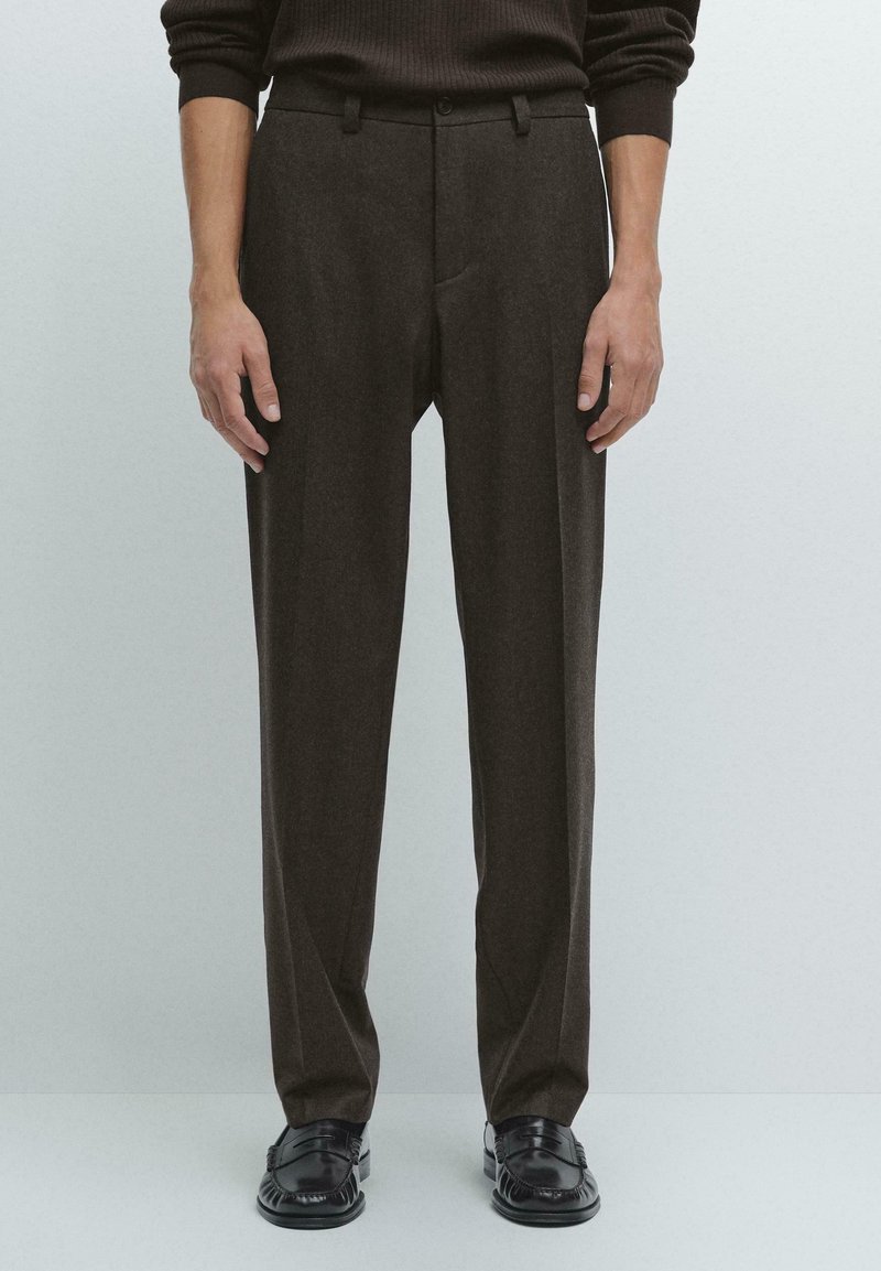 Man wearing dark brown tailored trousers and black leather loafers, standing against a plain light background.