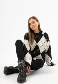 Un pull argyle noir et blanc avec un col côtelé, associé à un pantalon noir ajusté et des bottes à lacets noires épaisses avec un haut rembourré.