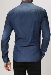 Tummansinisestä denim-kankaasta valmistettu paita, jossa on pitkät hihat, napilliset hihansuut ja klassinen kaulus. Sileä pinta ja räätälöity istuvuus, takana on selkäosa, jossa on hihanauhakuosi.