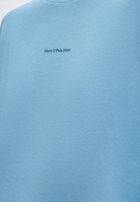 Φωτεινό γαλάζιο ύφασμα σε κοντινό πλάνο που δείχνει μικρό, κεντραρισμένο κεντημένο κείμενο "Marc O'Polo DENIM" με σκούρο μπλε κλωστή.