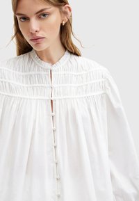 Blusa bianca realizzata in tessuto leggero, con un colletto alto arricciato, dettagli plissettati e chiusure con bottoni sul davanti.