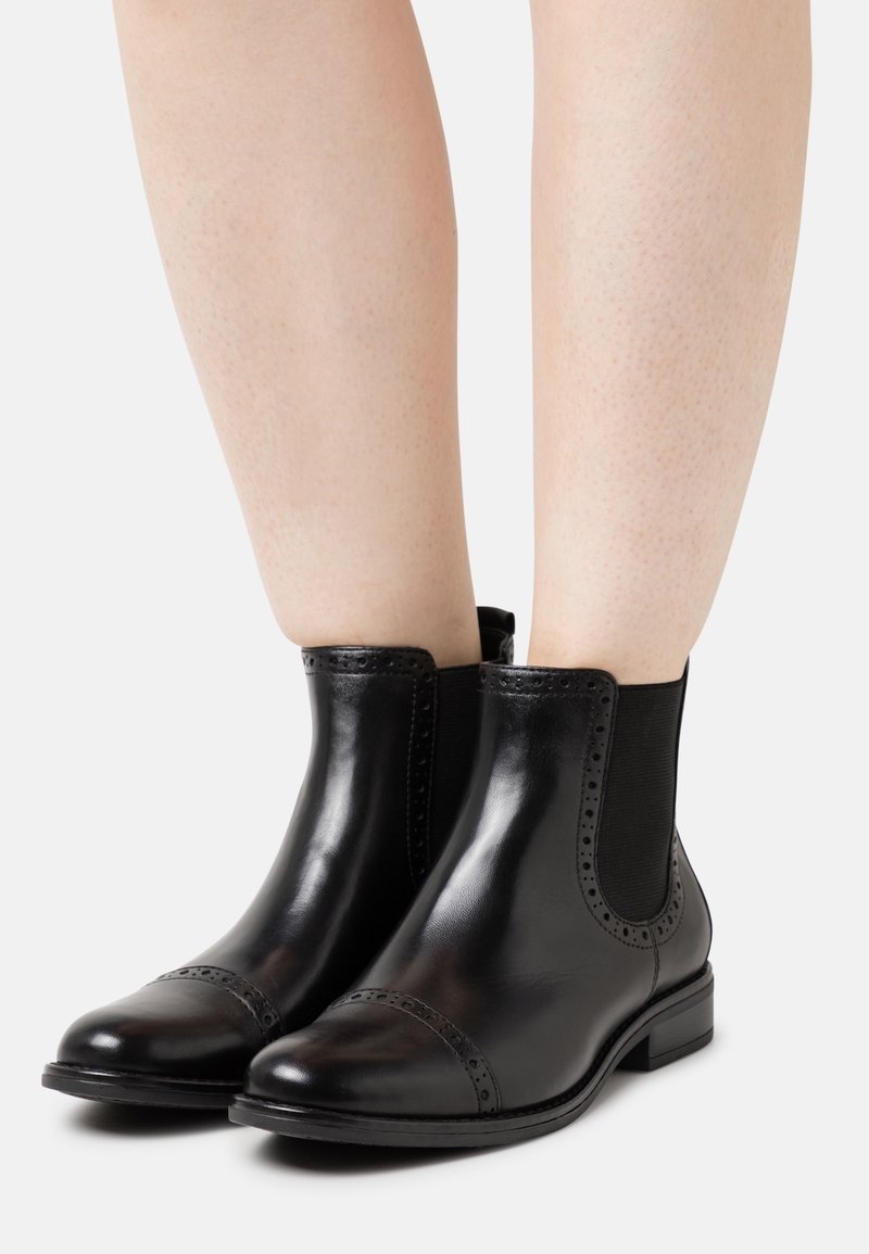 Bottines en cuir noir avec panneaux latéraux élastiques et détails à la brogue portées sur des jambes nues sur un fond clair uni.