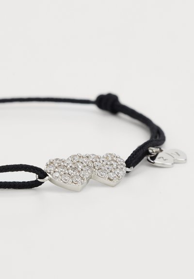 Brățară din cordon negru, cu două charm-uri în formă de inimă interconectate, decorate cu rhinestones clare, accente din metal argintiu și un mic charm în formă de inimă metalică.