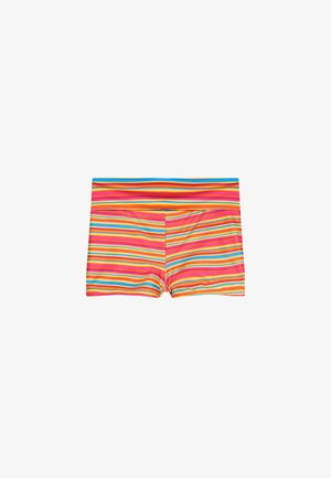 Junge Frau in buntem gestreiftem Bikini und beiger Kapuzenjacke steht an einem Strand mit felsigem Ufer und blauem Wasser im Hintergrund.