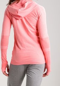 Person, der ein rosa Kapuzenshirt mit langen Ärmeln und graue Hose trägt, von hinten vor einem schlichten, hellen Hintergrund gezeigt.