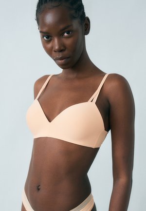 Γυναίκα που φοράει ένα απαλό σουτιέν σε γυμνό χρώμα με διπλές λεπτές τιράντες, σετ με το αντίστοιχο εσώρουχο, πάνω σε απλό ανοιχτό γκρι φόντο.