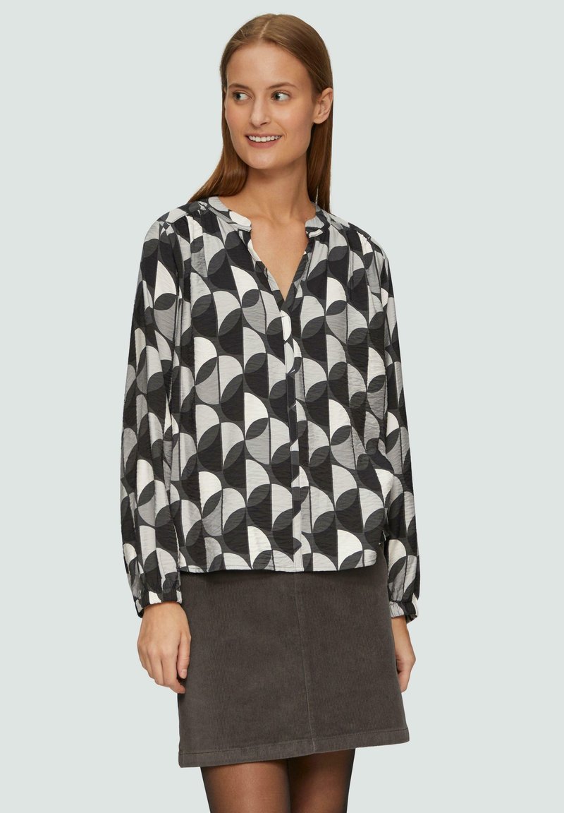 Blouse à motifs noir et blanc avec des formes géométriques, manches longues et patte de boutonnage, associée à une jupe en velours côtelé gris.