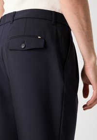 Dark navy trousers made of smooth fabric, featuring a back pocket with a button closure and a subtle decorative accent on the pocket.