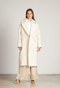Abrigo largo de marfil con bordado floral, diseño de doble botonadura, amplios solapas y dos bolsillos exteriores, usado sobre un atuendo de color claro.