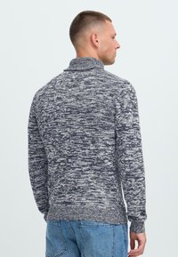 Moški s kratkimi lasmi, oblečen v melirano sivo-belo pleteno jopico in modre jeans hlače, obrnjena stran od preproste svetle ozadja.
