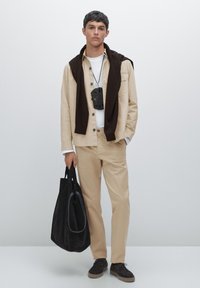 Light beige jacket and pants, white shirt underneath, brown sweater draped over shoulders, black bag held in hand, dark shoes.