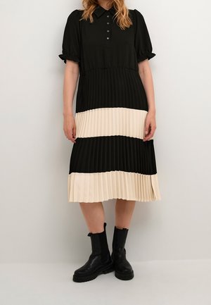 Donna che indossa un vestito midi plissettato bicolore nero e crema con maniche corte a sbuffo e stivali neri robusti alla caviglia.