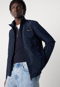 Veste zippée bleu marine en matériau lisse et résistant à l'eau. Dotée d'un col haut et d'un petit logo Calvin Klein sur la poitrine.