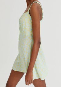 Robe florale en vert pastel avec un motif de fleurs jaunes. Présente un col à volants et des bretelles fines. Tissu léger, forme A-line.