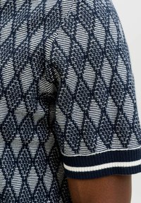 Short-sleeve knitted sweater with navy and white diamond pattern and striped cuff on a dark-skinned arm.
