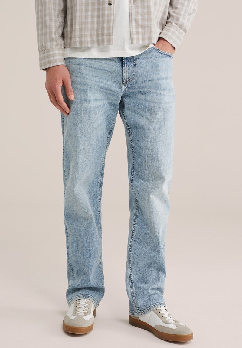 Uomo che indossa jeans a gamba dritta azzurri, sneaker beige, una camicia bianca e una giacca a quadri beige, in piedi con una mano in tasca.