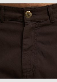 Bruine katoenen broek met een rits en knoopsluiting. Zichtbaar logo op de knoop, met stiksels langs de tailleband en zakken.