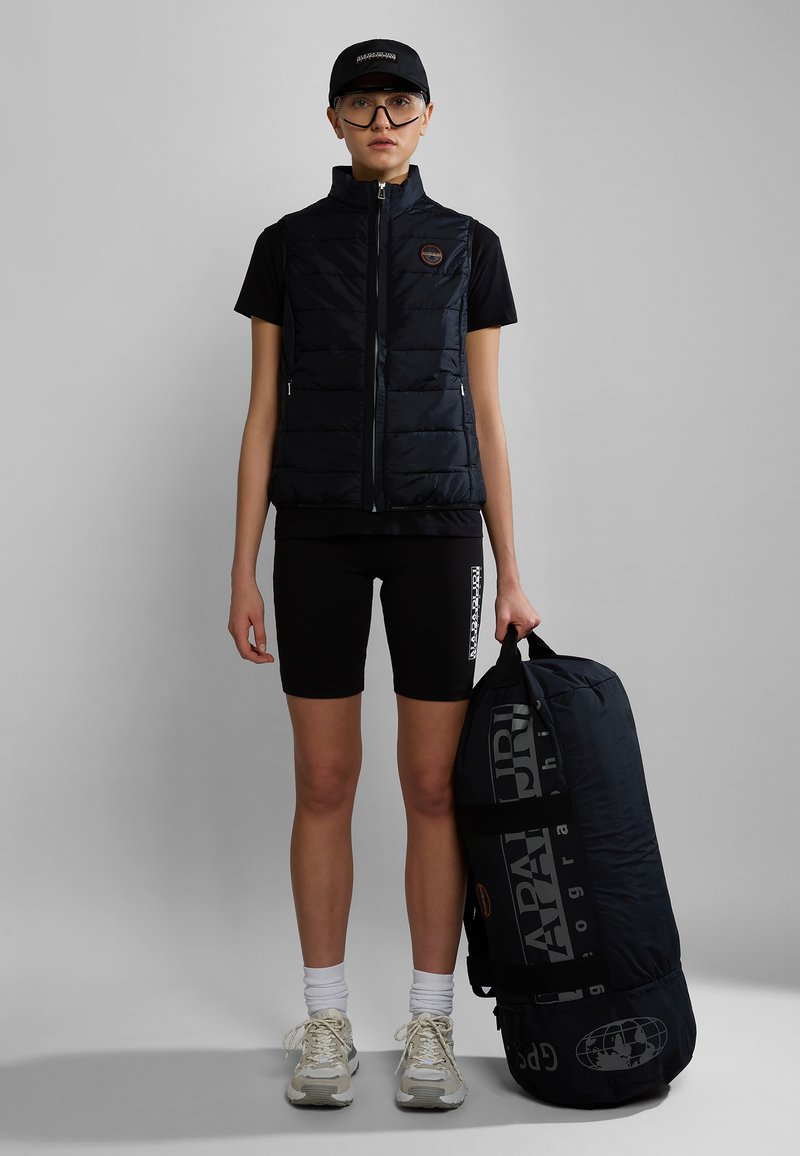 Czarna, insulated kamizelka z wysokim kołnierzem, zestawiona z czarną koszulą i szortami. Trzyma dużą, czarną torbę podróżną z białymi akcentami graficznymi.