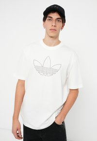 Balta kokvilnas T-kreklu ar īsām piedurknēm, ar pelēku punktotu Adidas emblēmu priekšpusē. Modelis valkā melnas bikses un melnu cepuri.