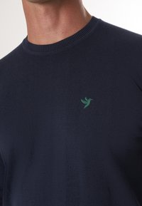 Marineblauwe gebreide trui met een ronde hals en een klein groen kolibrilog op de borst. De stof heeft een gladde textuur.