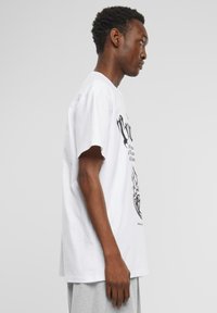 Hvid bomuld t-shirt med løs pasform, featuring sorte grafiske tryk af flammer og tekst på fronten. Korte ærmer og rund halsudskæring.