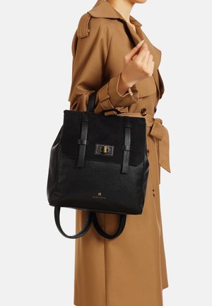 Borsa tote in pelle nera e suede dalla forma strutturata, con dettagli in hardware dorato e manici regolabili, tenuta da una persona che indossa un cappotto beige.