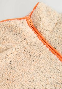 Tissu polaire beige doux avec des taches noires, fermeture éclair orange vif et bordure sur un vêtement à col montant.