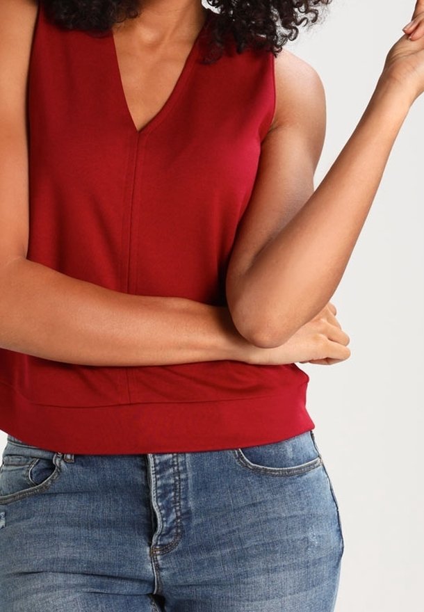 Červený top bez rukávů s výstřihem do V, vyrobený z hladkého materiálu. Spodní lem je mírně zakřivený. V kombinaci s modrými džíny.