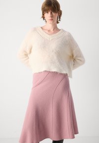 Pulover scurt, fuzos, de culoare crem, cu decolteu în formă de V; asortat cu o fustă roz, de lungime până la genunchi, în stil A, care are un detaliu cu cusătură diagonală.