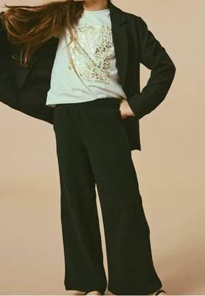 Mustat leveälahkeiset housut, joissa on teksturoidu pinta, yhdistetty vaalean väriseen graafiseen t-paitaan, jossa on leopardi-kuvio ja metallinhohtoiset yksityiskohdat, mustan bleiserin alla.