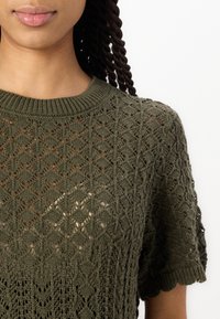 Nahaufnahme einer Person, die ein olivgrünes gestricktes Oberteil mit einem komplexen Diamant- und Blumenmuster trägt, wobei die Details von Hals und Schultern zu sehen sind.