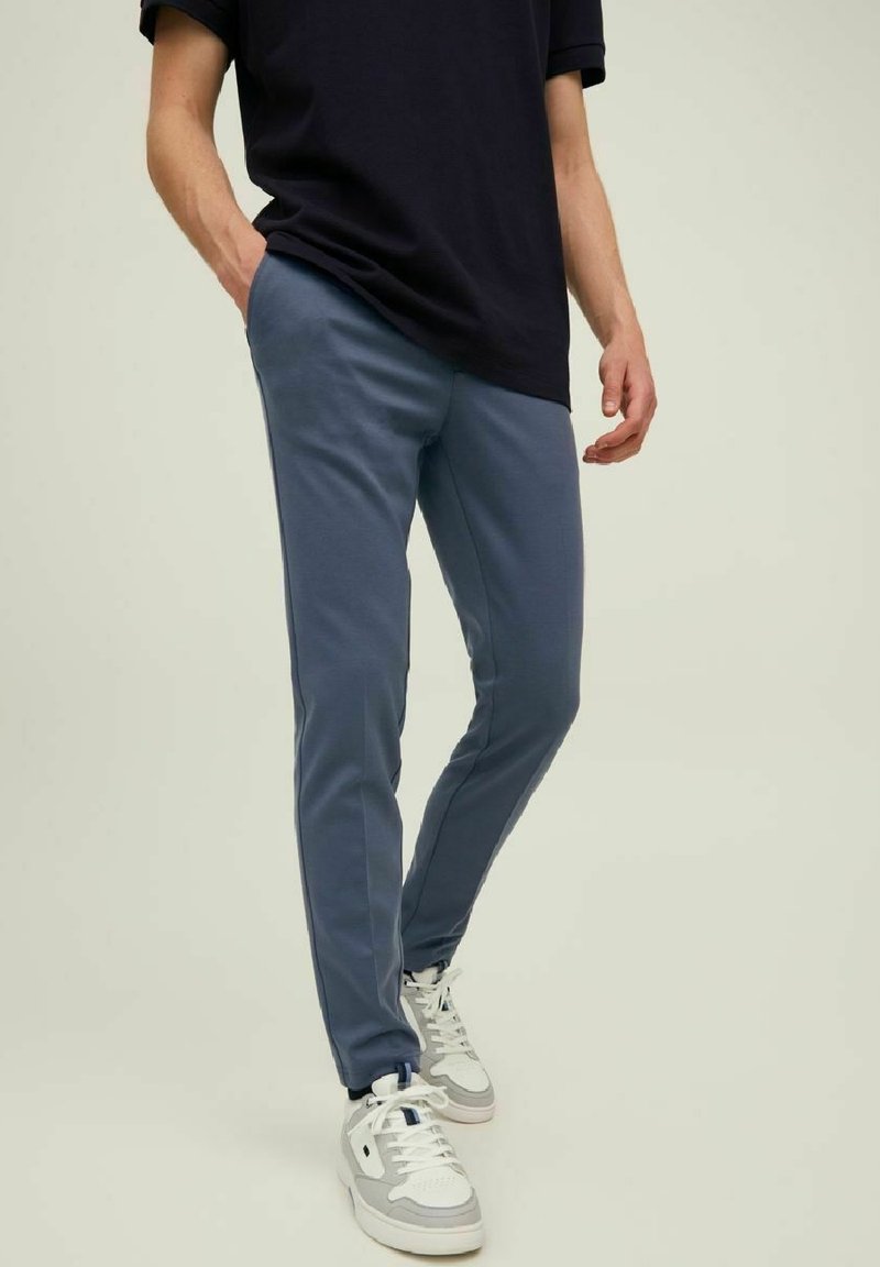 Hombre vestido con camiseta azul marino, pantalones ajustados azules, zapatillas blancas y grises, con una mano en el bolsillo, de pie sobre un fondo claro.