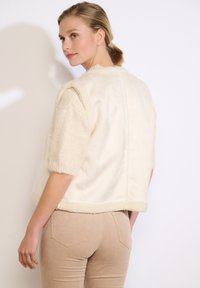 Crème-kleurige faux fur jas met korte, gestructureerde mouwen en een recht gesneden ontwerp. Voorzien van een zachte, pluche afwerking en randaccenten.